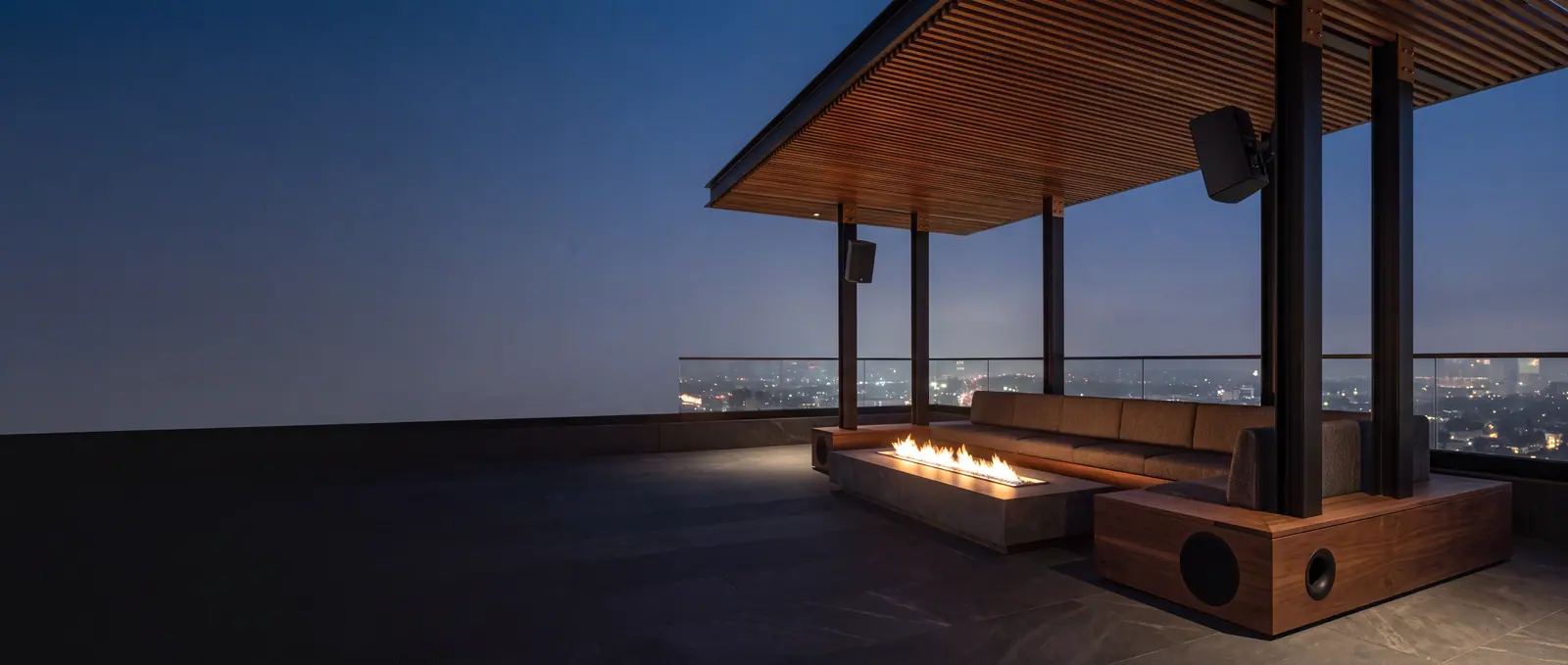 Design concept: a Los Angeles rooftop terrace at blue hour with compact loudspeakers mounted to the pergola structure and a subwoofer integrated into the bench plinth.